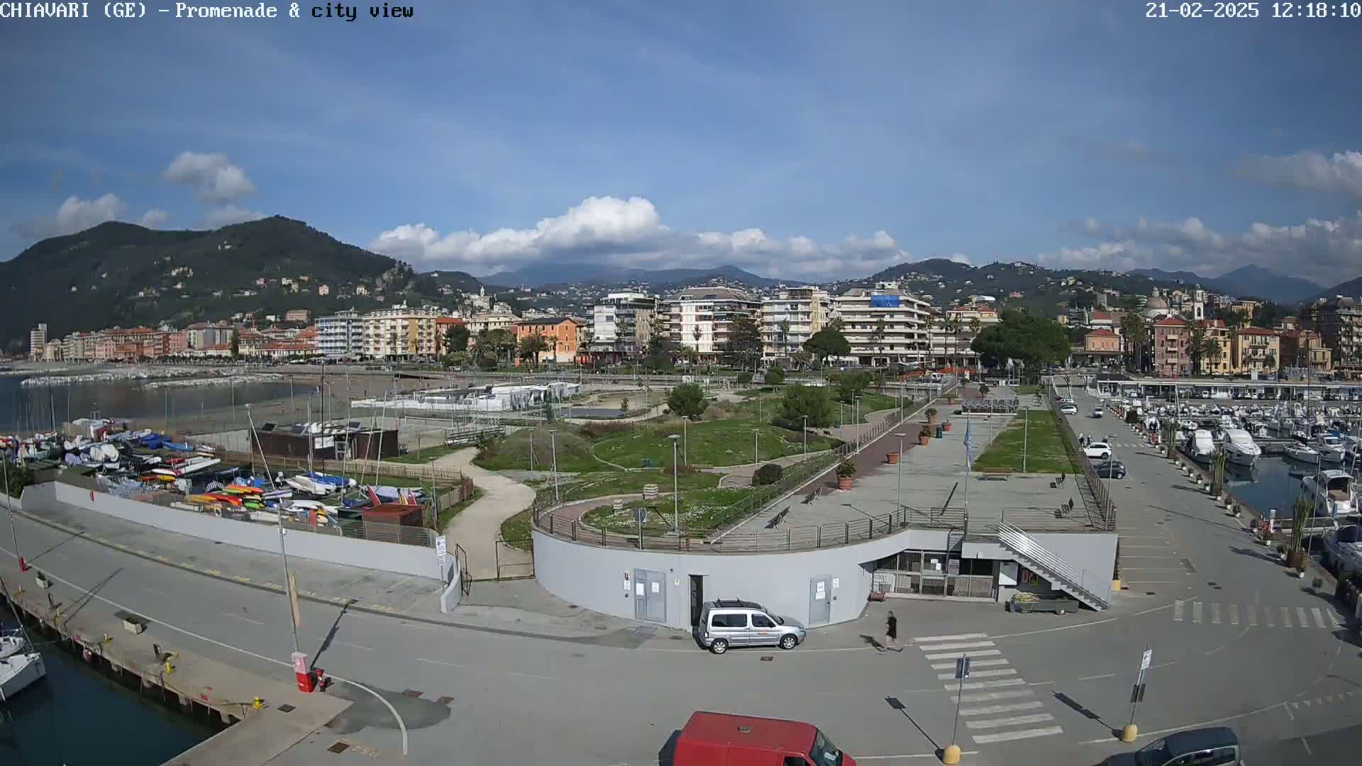 A mostly sunny day overlooking a coastal town's harbor, promenade, and a park with numerous boats docked, and a few cars parked.