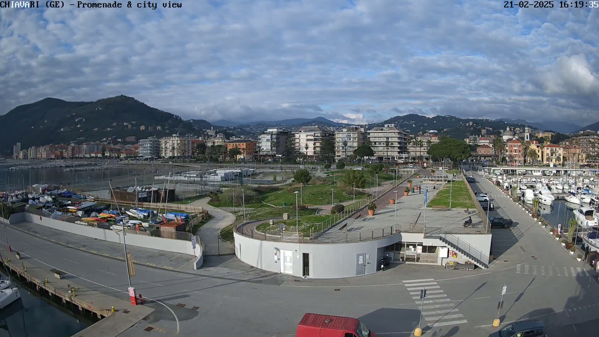 A harborside promenade with a marina, green spaces, and city buildings in the background under a partly cloudy sky.