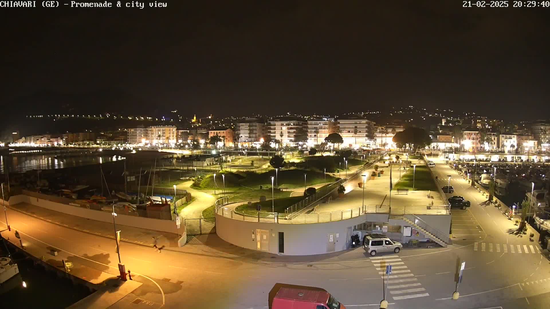 A nighttime aerial view of a harbor and city, showing a promenade, park, and boats docked at the waterfront under clear skies.