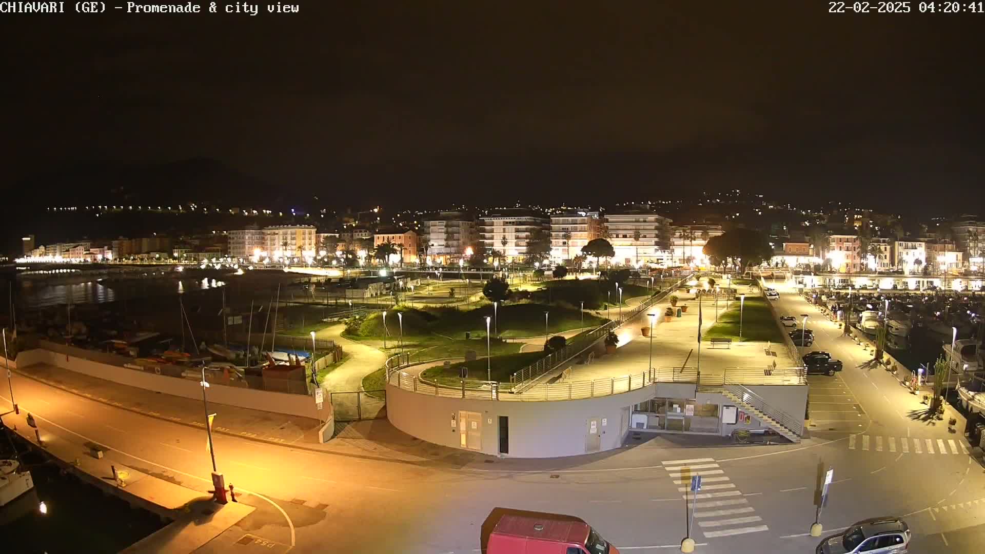 A nighttime aerial view of a city's illuminated waterfront promenade and harbor, featuring a park with winding pathways and boats docked along the shoreline.