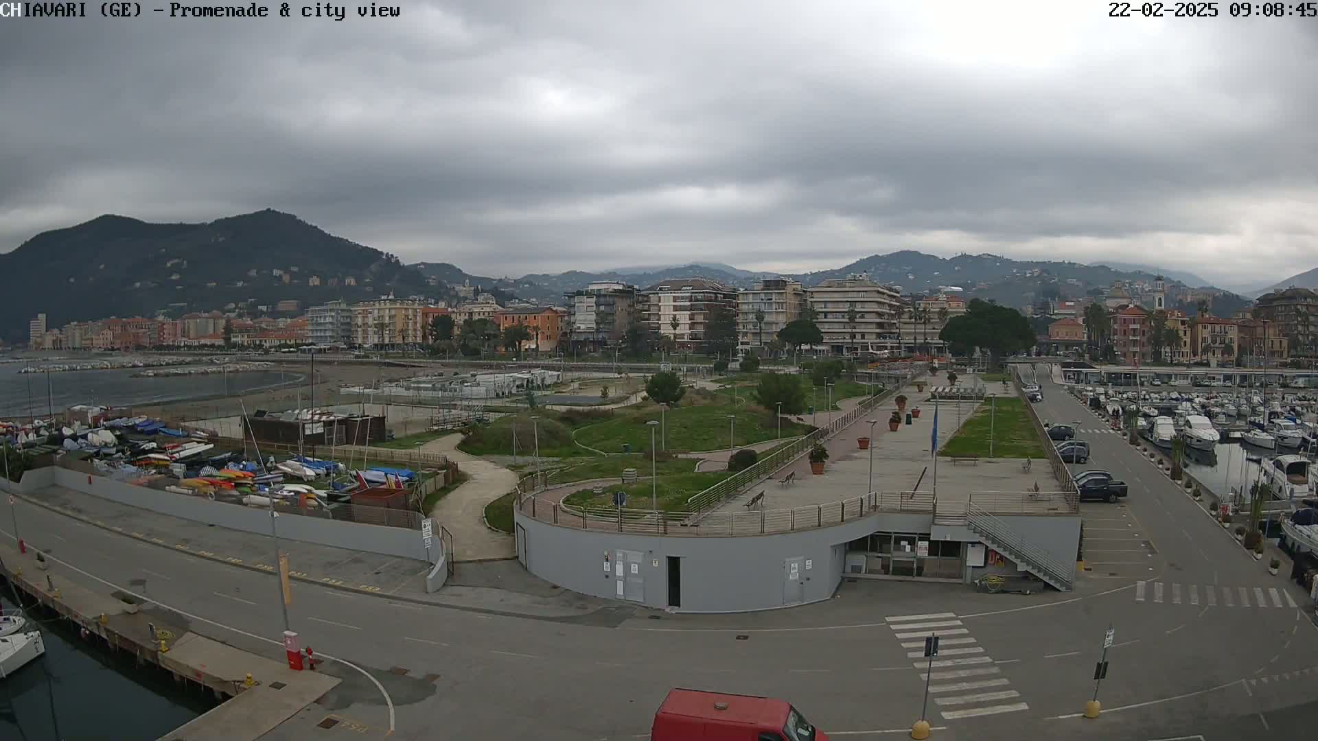 A coastal city scene under an overcast sky shows a promenade, marina with boats, and apartment buildings nestled against a mountain backdrop.