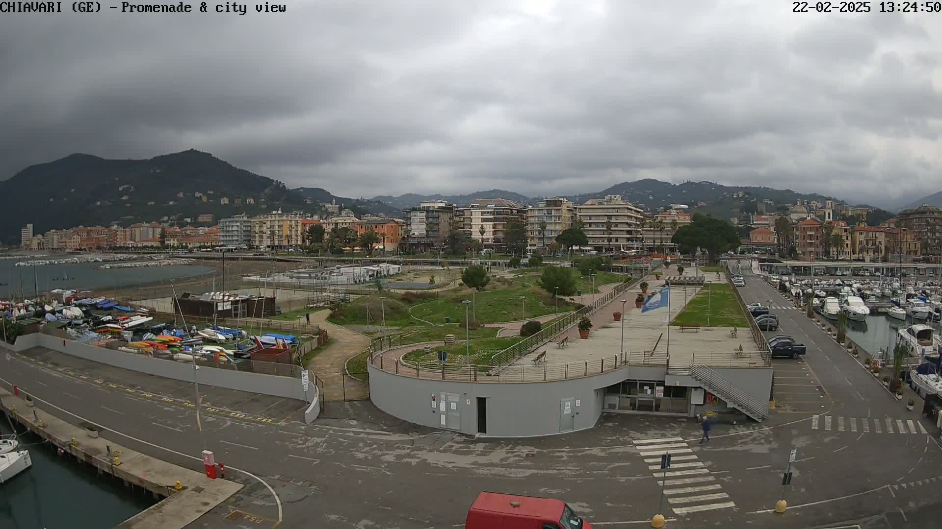 An overcast day reveals a coastal city with a harbor filled with boats, a promenade with green spaces, and buildings stretching along a hillside.