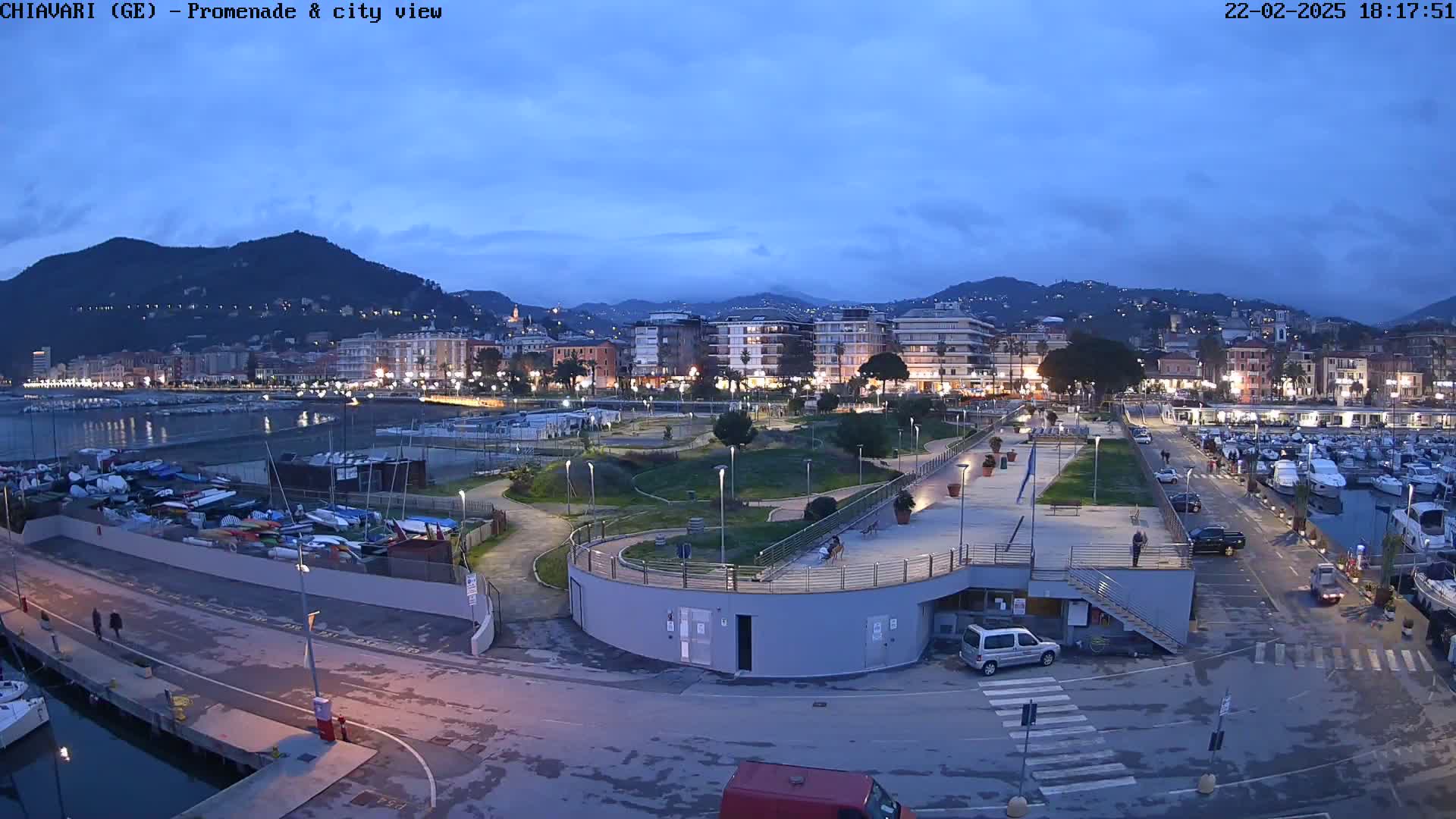 A nighttime view of a harbor, marina, and city under a twilight sky, showing a waterfront park with boats, cars, and buildings.