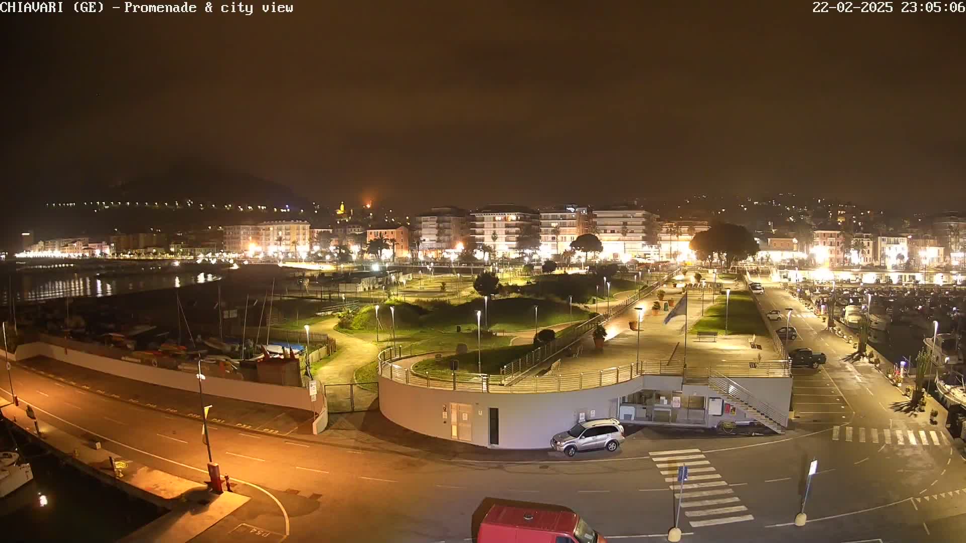 A nighttime view of a brightly lit coastal town's promenade and harbor, featuring a park, parked cars, and boats.