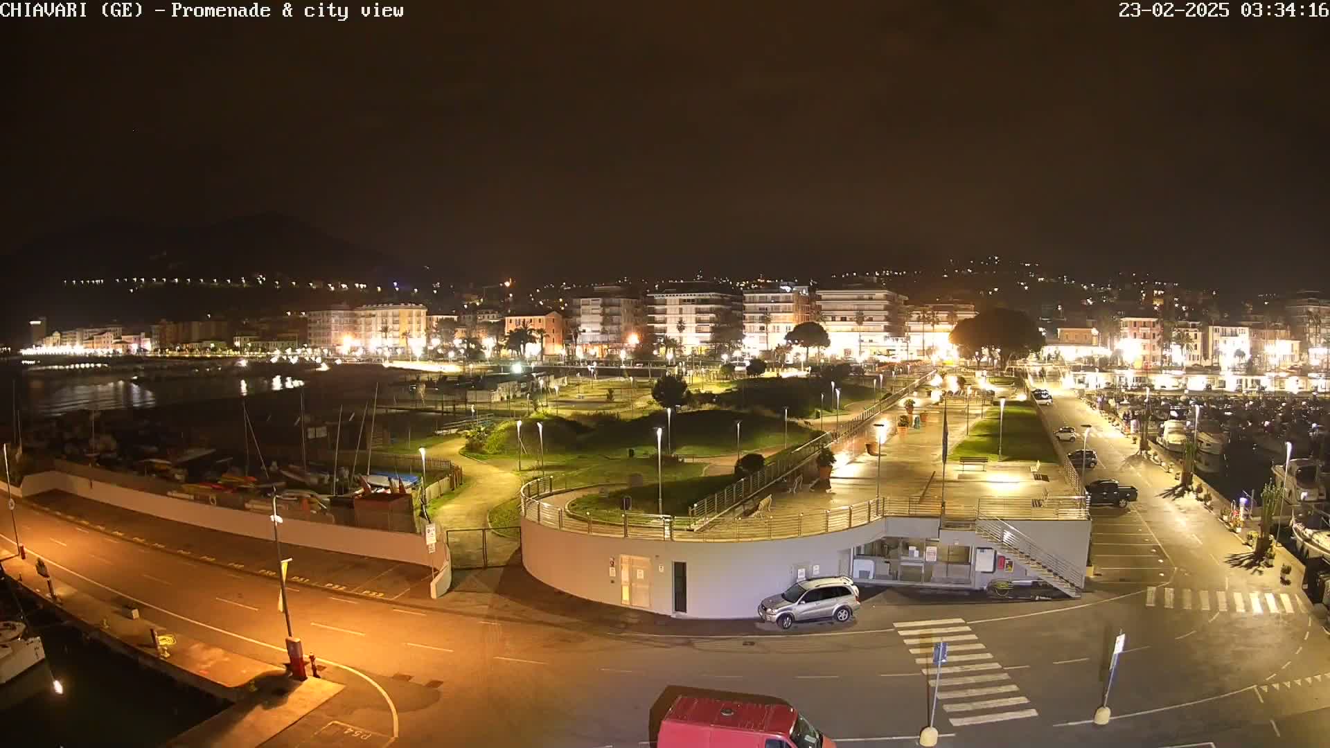 A nighttime aerial view of a coastal city's illuminated promenade and harbor, with boats moored and cars parked, under a dark, clear sky.