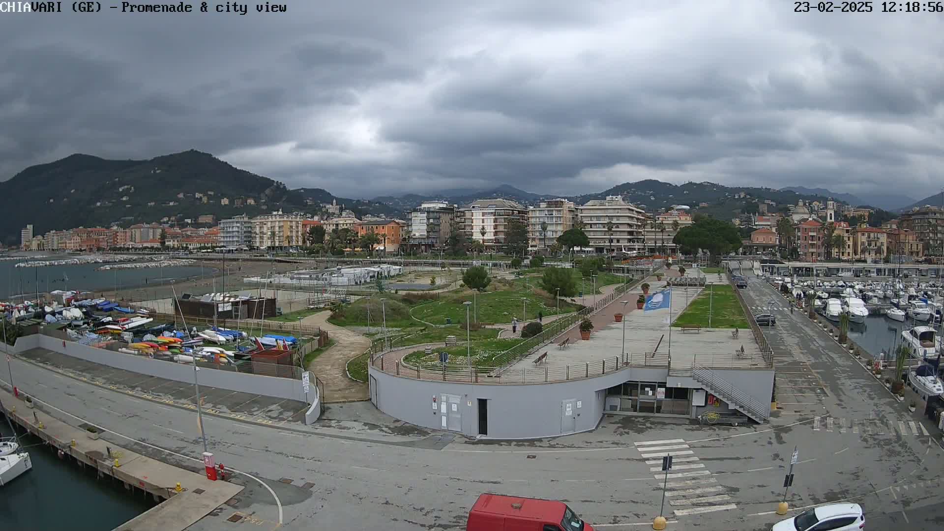 A coastal city scene under a cloudy sky shows a harbor filled with boats, a park, and buildings along a waterfront promenade.