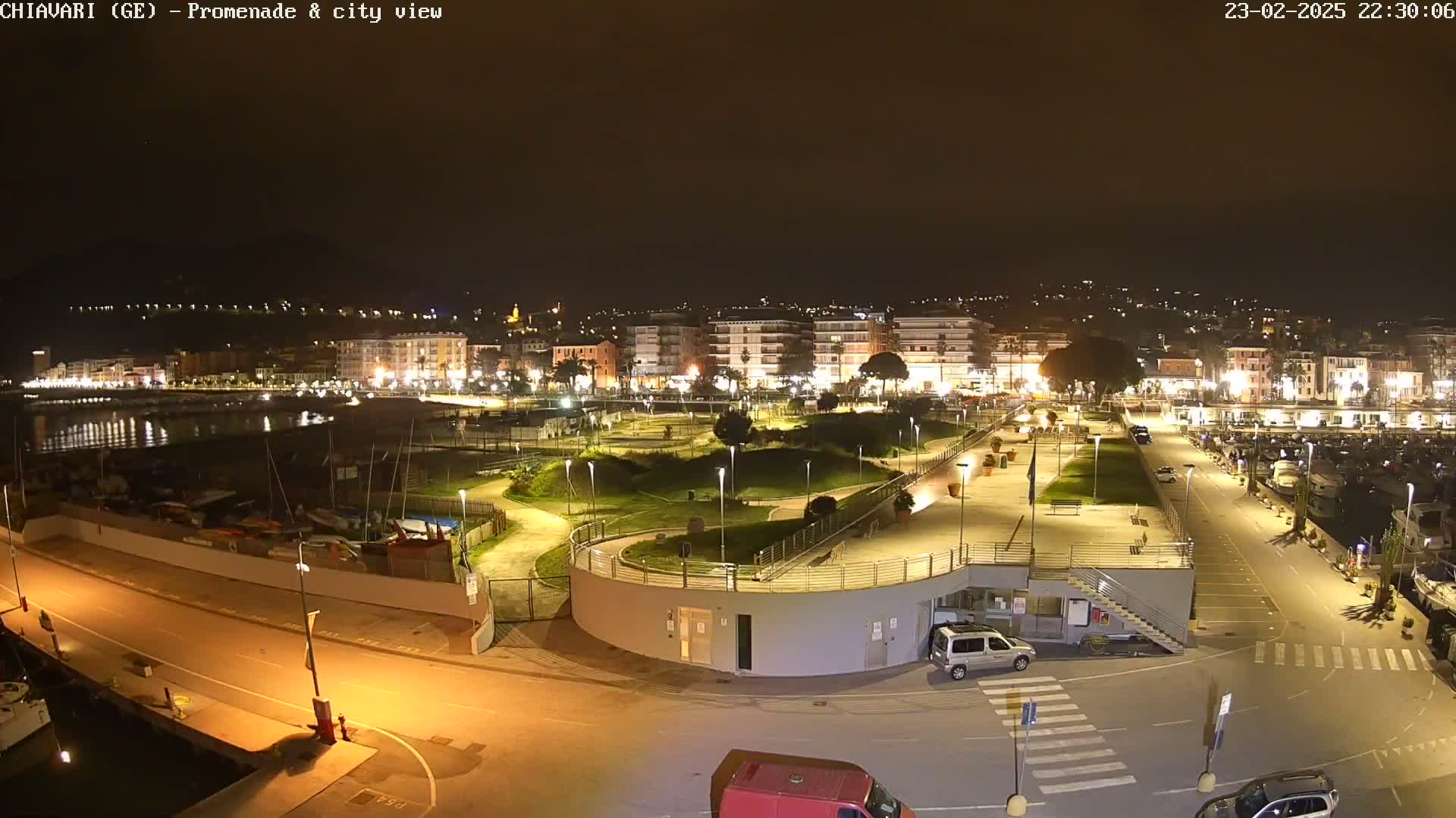 A nighttime view of a harbor, promenade, and city, brightly lit with several parked cars and boats visible in the marina.