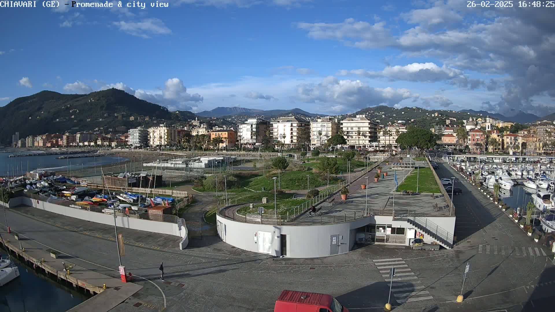 A waterfront promenade with a marina, green spaces, and a town in the background under a partly cloudy sky.