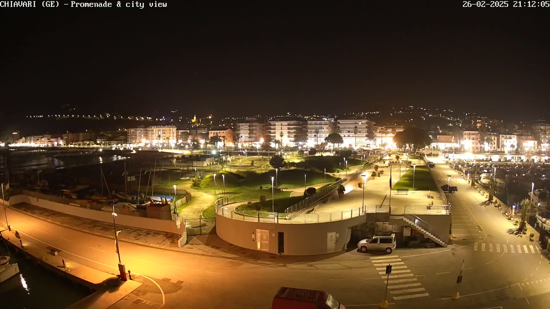 A nighttime view of a city's waterfront promenade and harbor, showing a well-lit park and marina with boats, and buildings in the background.