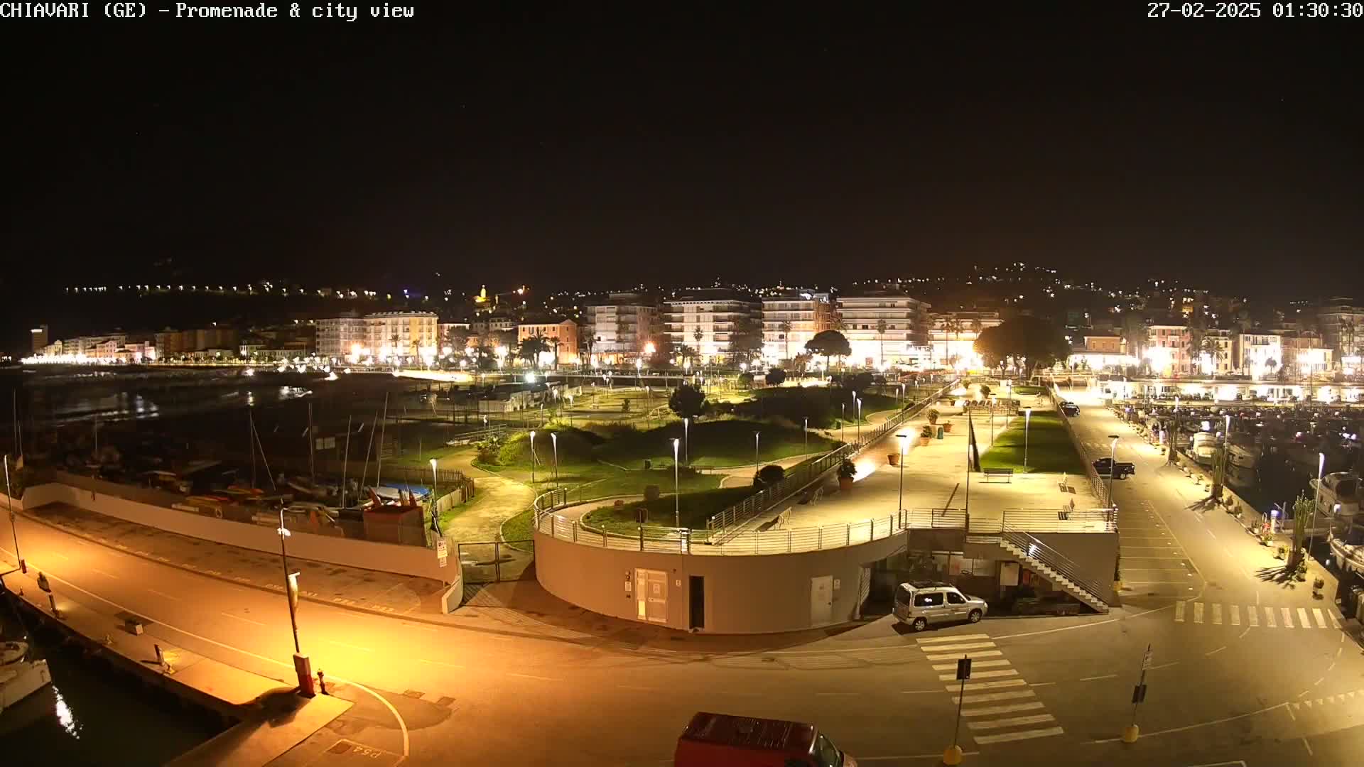 A nighttime aerial view of a city's waterfront promenade and marina, illuminated by streetlights, with a calm body of water and parked boats visible.