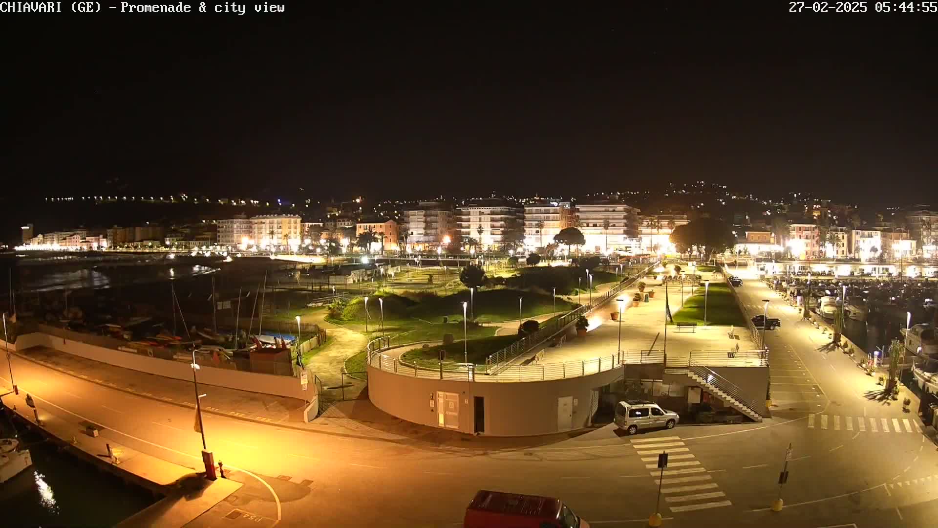 A nighttime view of a city's illuminated waterfront promenade and harbor, featuring a landscaped park, parked cars, and boats in the marina.