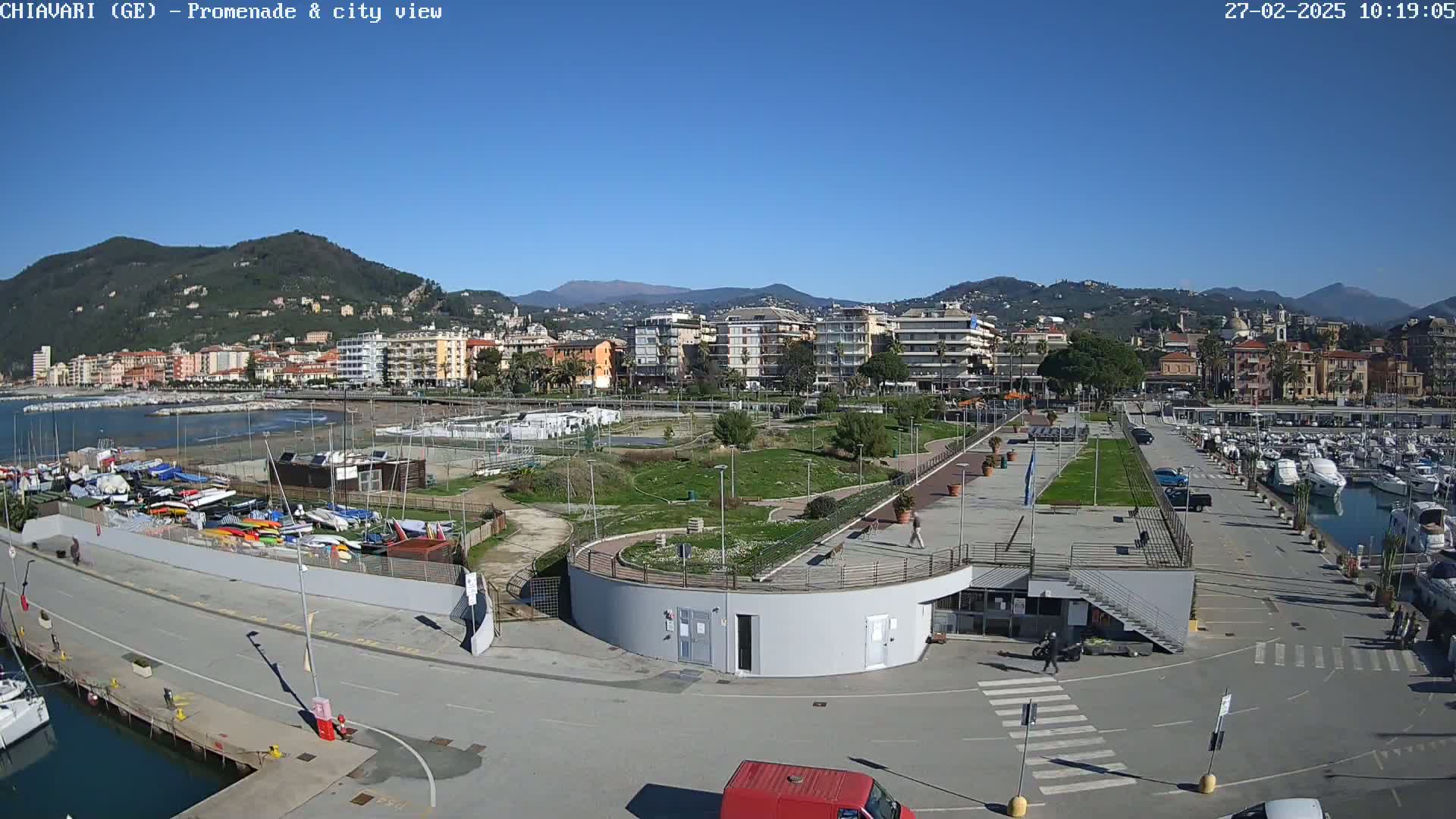 A seaside town's promenade and harbor are shown on a sunny day, with boats docked and a grassy area separating the walkway from the buildings and water.