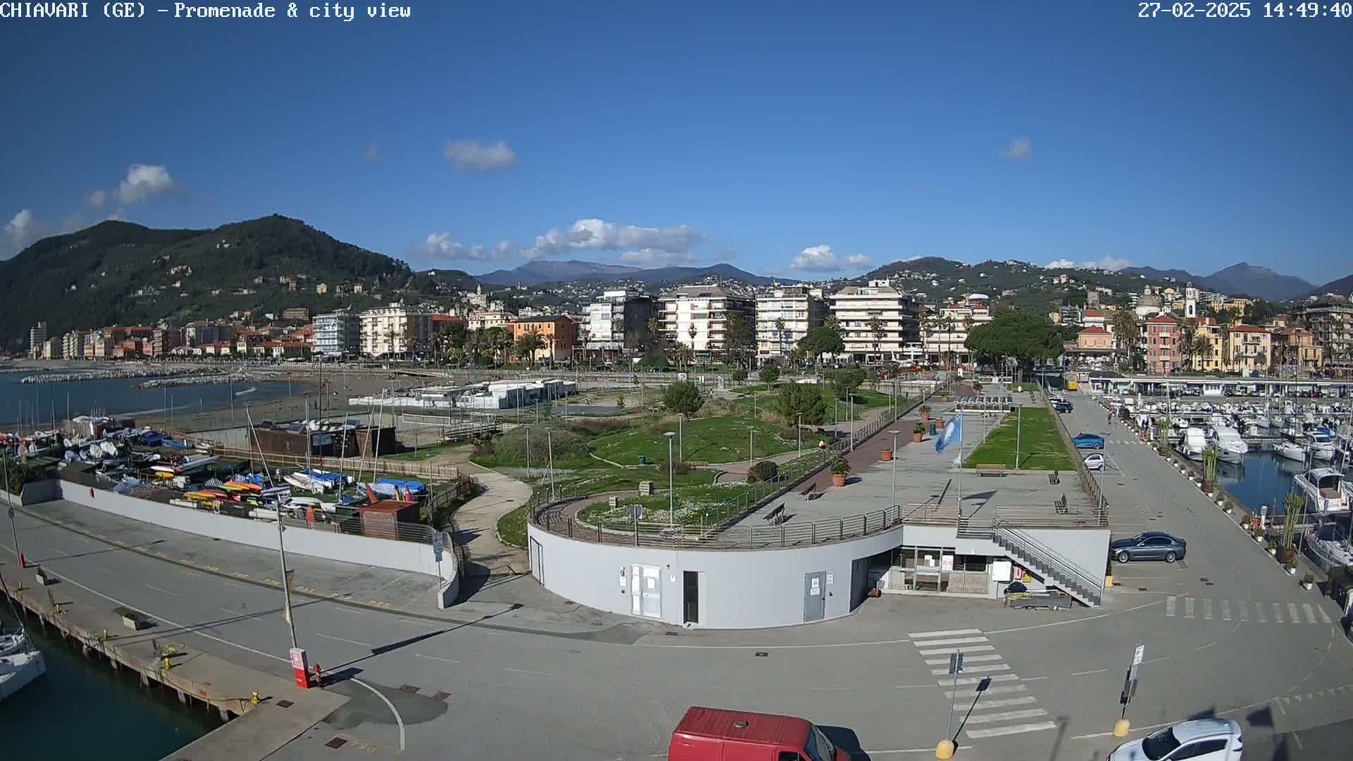 An aerial view of a coastal town on a sunny day, showing a harbor filled with boats, a promenade with a park, and buildings extending into the hills.
