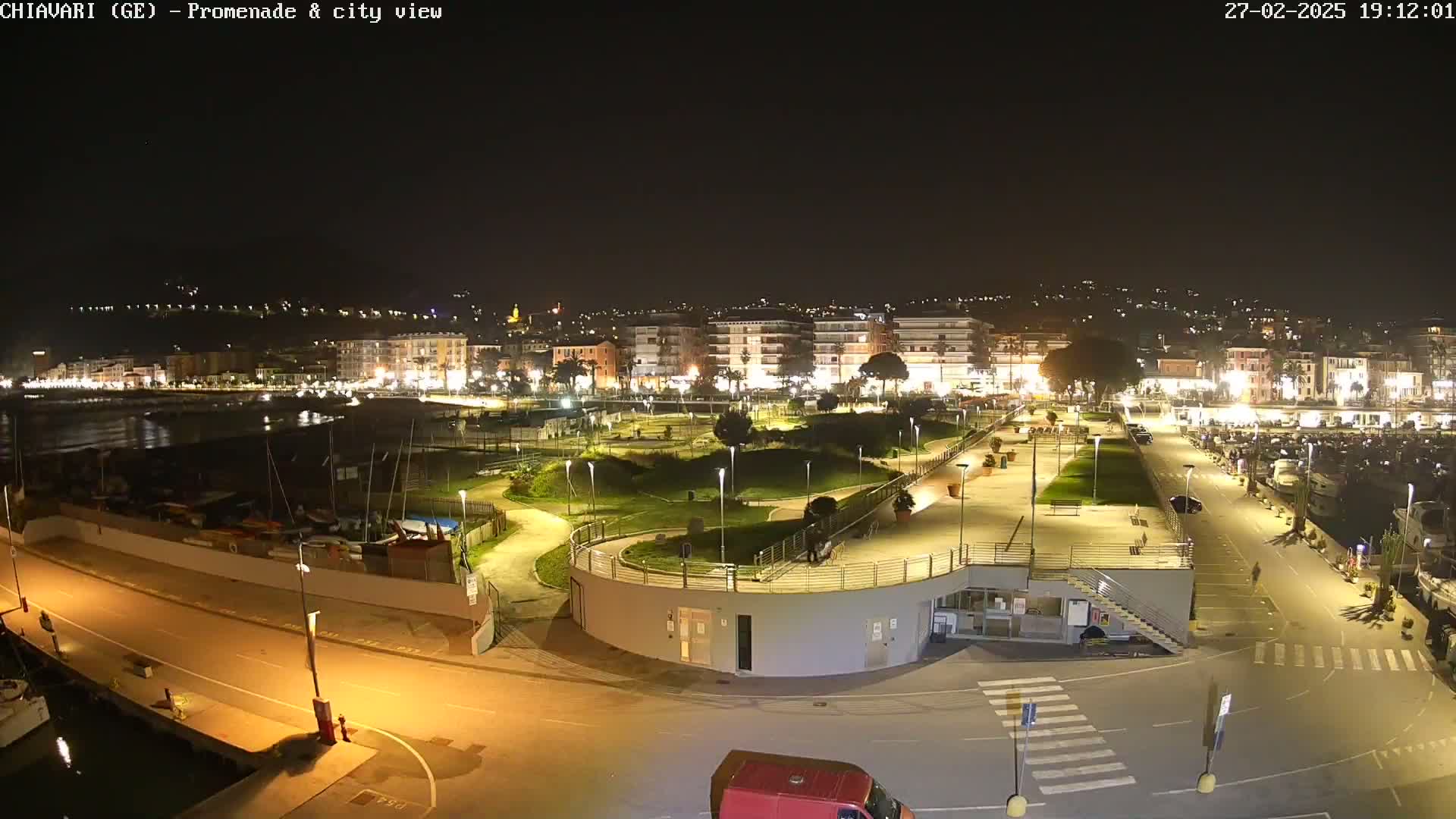 A nighttime view of a city's waterfront promenade and marina, with well-lit buildings and landscaping, under a clear, dark sky.