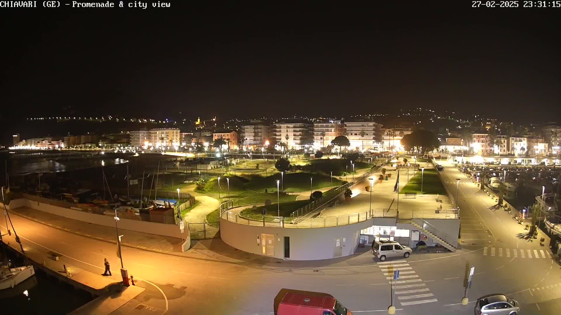 A nighttime view of a brightly lit seaside promenade and city, with a calm body of water and numerous boats visible in the harbor.