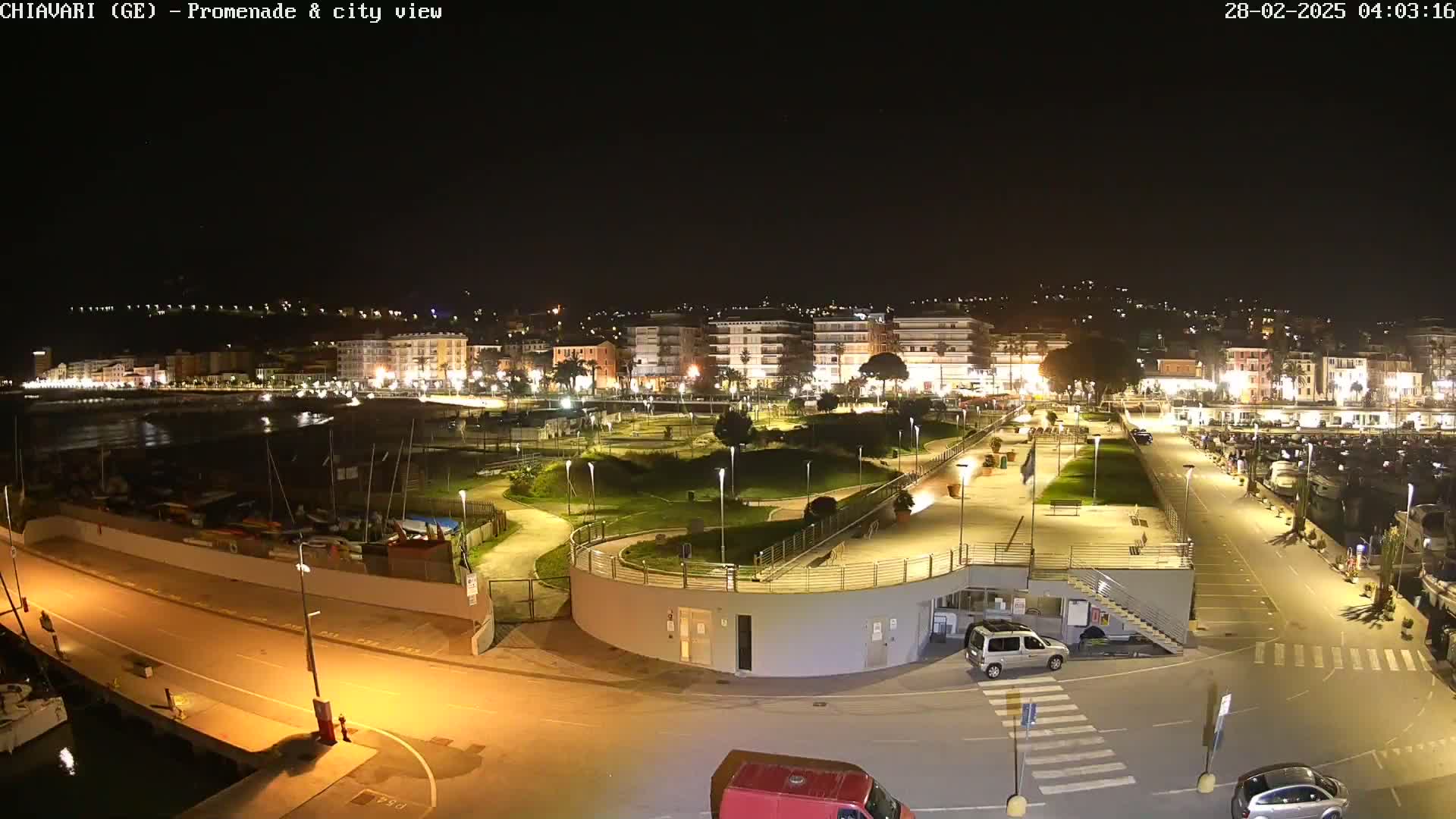 A nighttime, aerial view of a city's waterfront promenade and marina, brightly lit, with a small park and several vehicles visible.