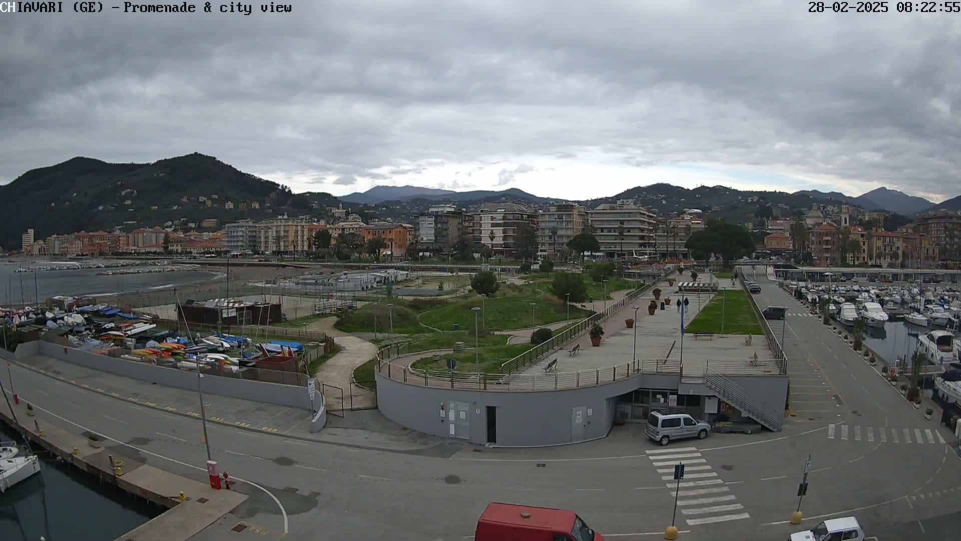 An aerial view shows a harbor with numerous boats, a promenade with green space, and a town nestled against a mountain range under a cloudy sky.