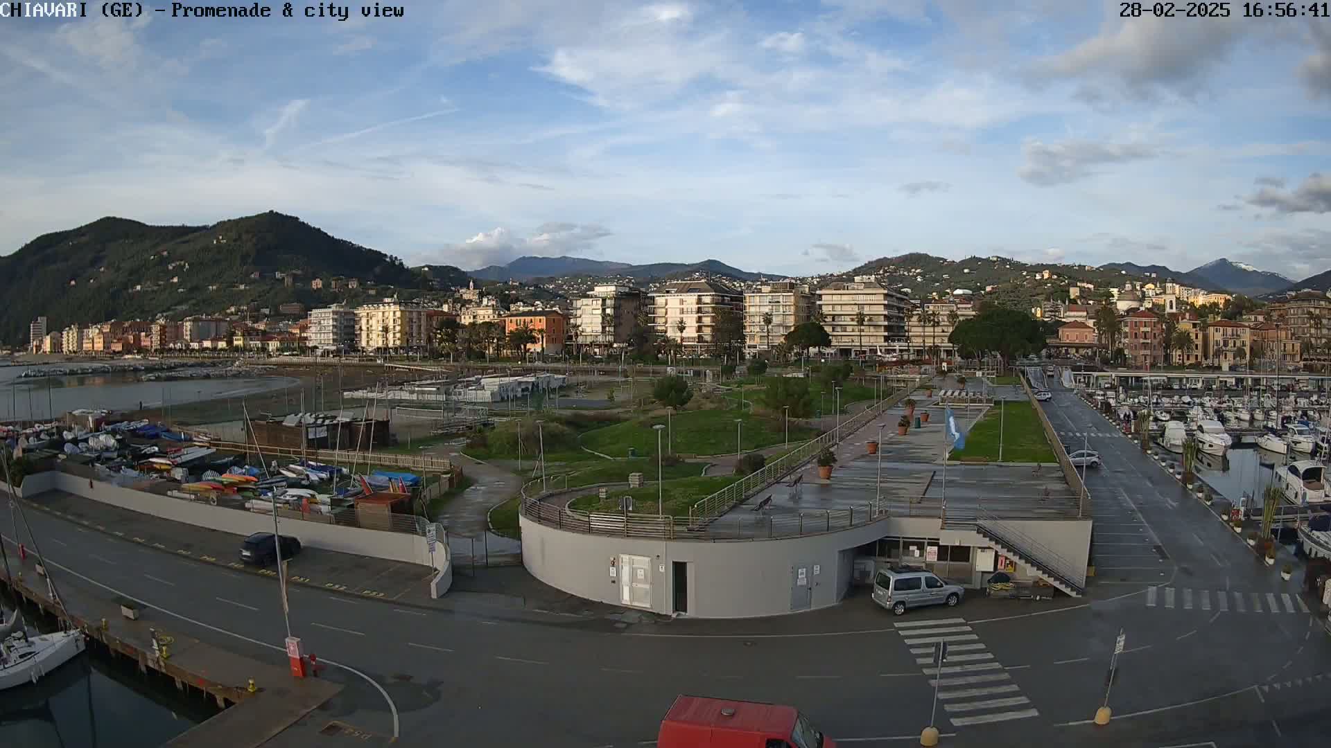 An aerial view of a coastal city with a marina, park, and buildings under a partly cloudy sky.