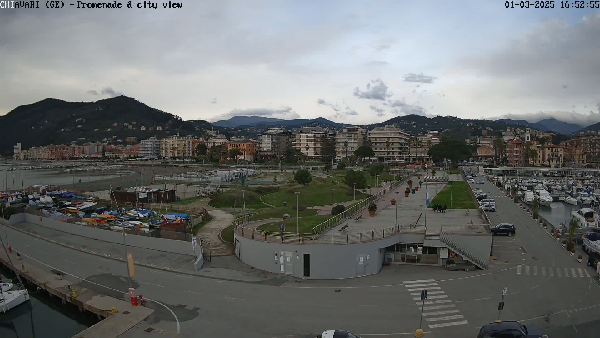 An overcast day reveals a coastal city scene with a marina filled with boats, a promenade, a grassy park, and multi-story buildings nestled against a backdrop of mountains.