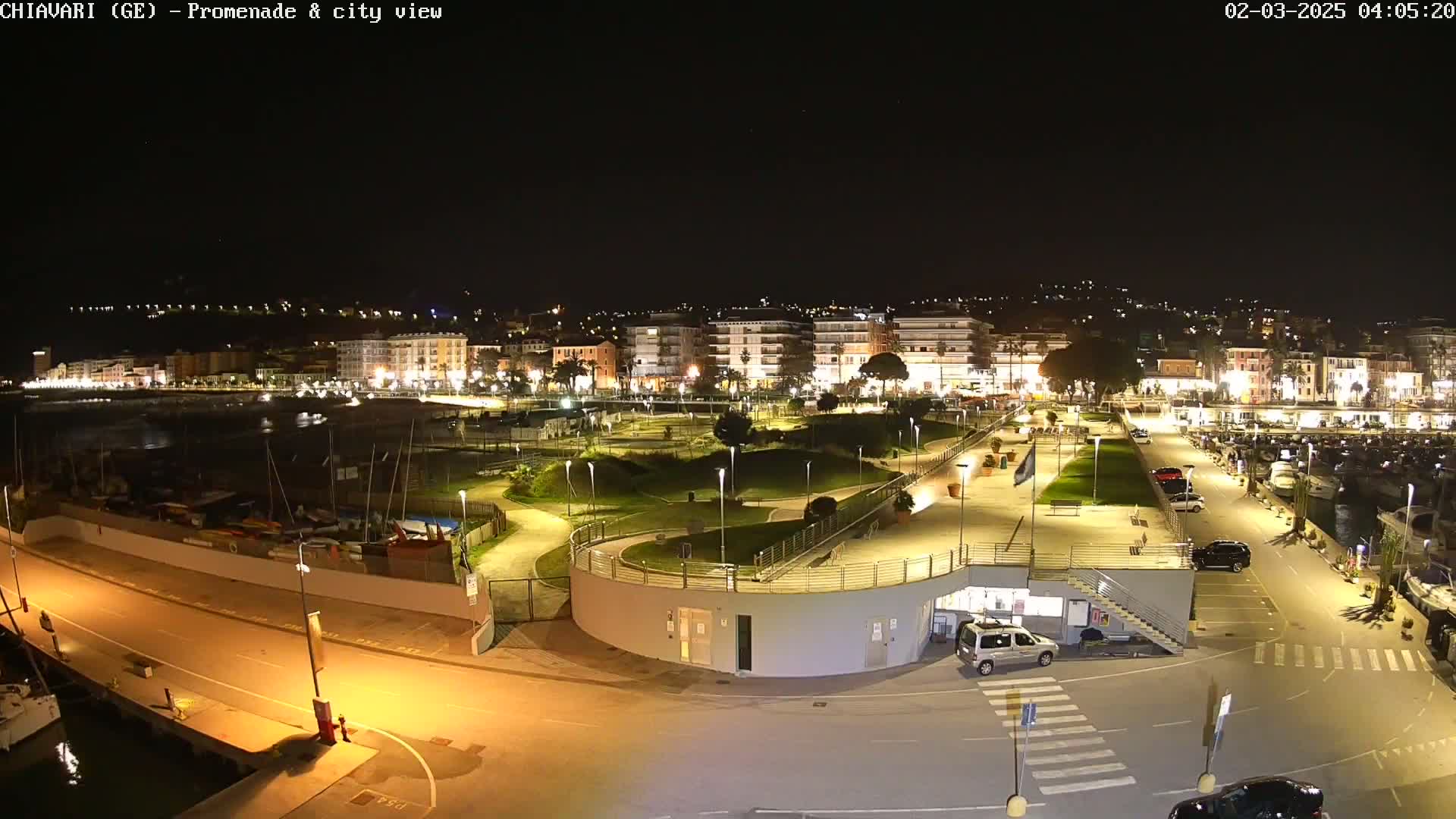 A nighttime, illuminated view of a waterfront promenade and city, featuring a marina with boats, a park-like area, and buildings along the shore.
