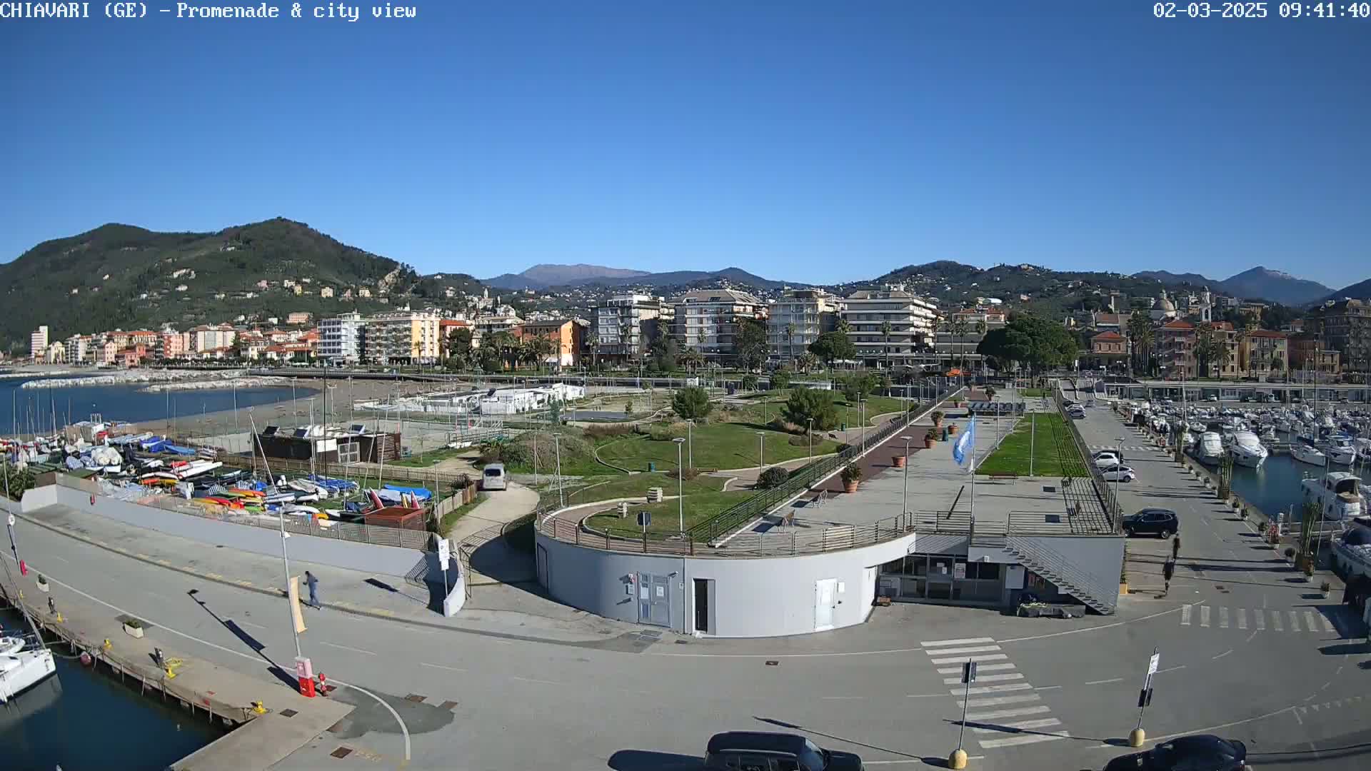 A waterfront promenade with a marina and a town nestled against a hillside under a clear blue sky.