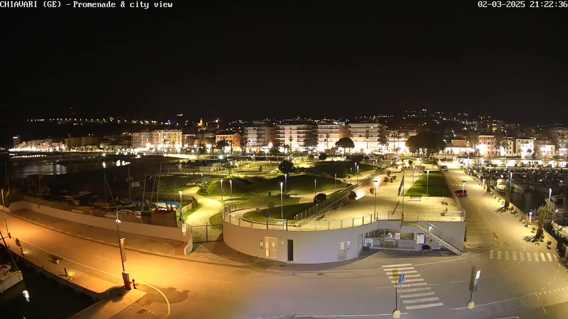 A nighttime view of a brightly lit seaside promenade and city, with a marina and park visible.