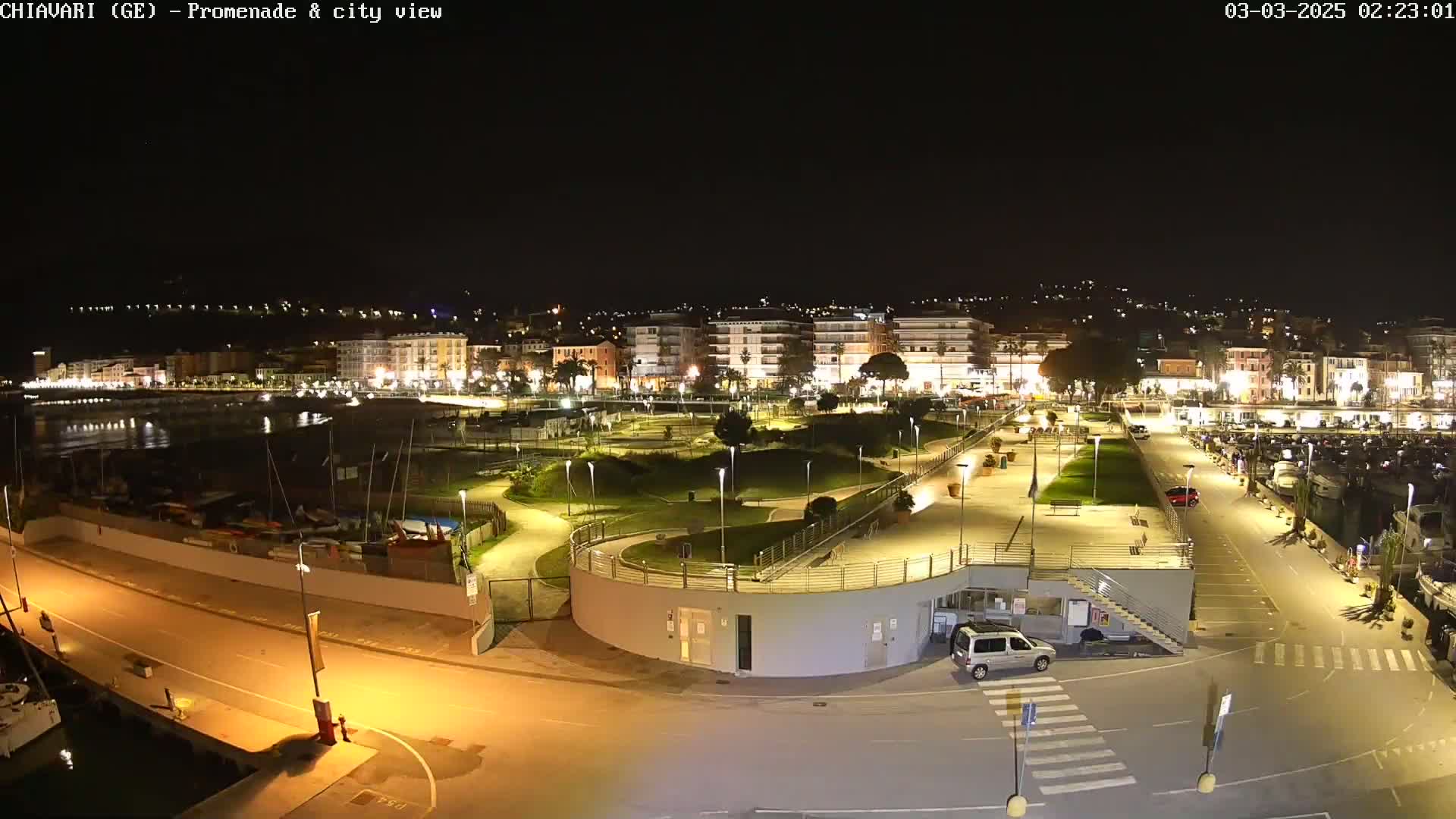 A nighttime, brightly lit waterfront promenade and city view shows a harbor with boats, a park-like area, and buildings along the shoreline.