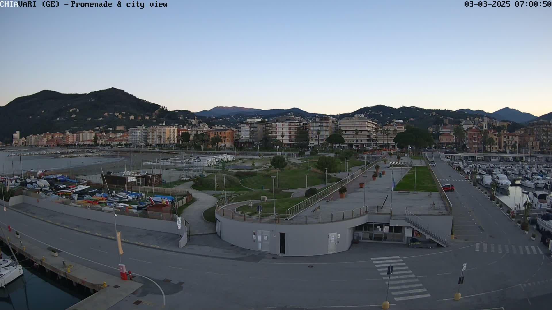 A waterfront promenade with a marina, a grassy park, and a city with mountains in the background under a clear twilight sky.