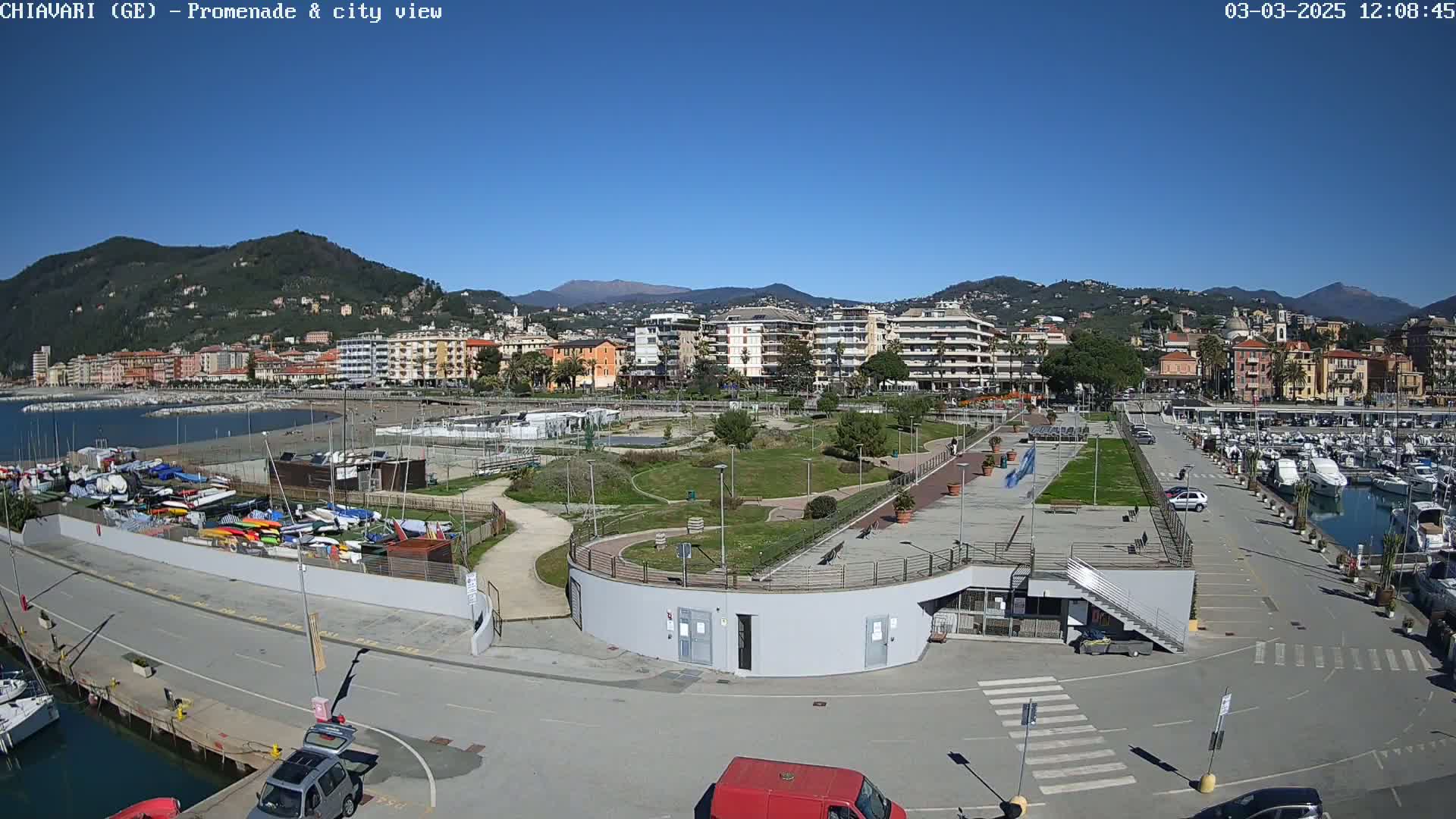 A waterfront promenade with a marina, a park, and a city in the background under a clear blue sky.