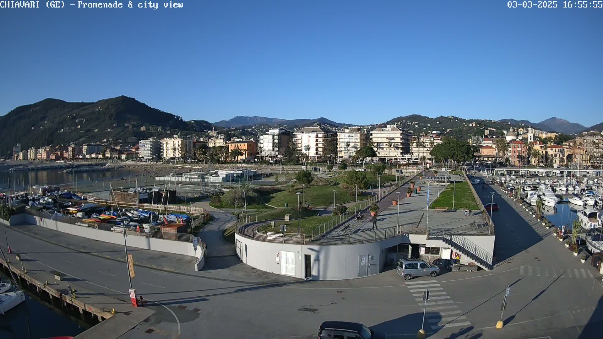 An aerial view shows a seaside town on a sunny day, with a harbor full of boats, a promenade, and green spaces separating the waterfront from residential buildings and hills in the background.