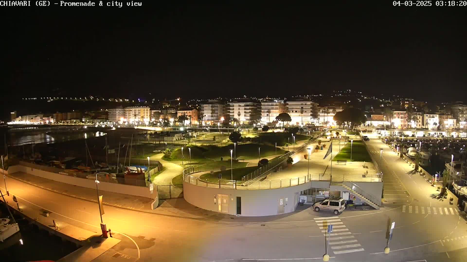 A nighttime aerial view of a waterfront promenade and city, illuminated by streetlights, with a calm body of water and moored boats visible.
