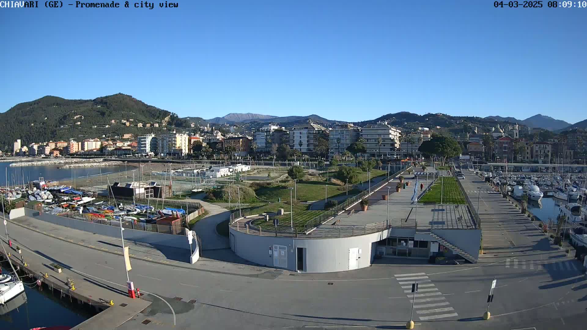 A clear, sunny day overlooks a harbor filled with boats, a promenade, a park, and a city nestled against mountains in the background.
