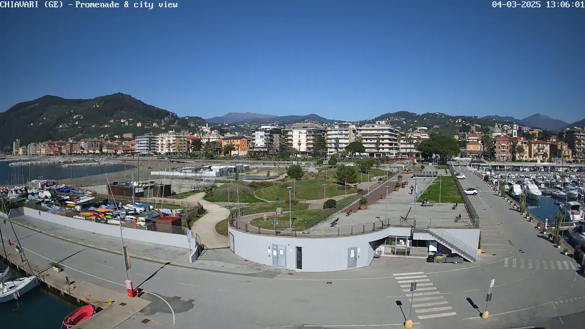A seaside town with a marina, a promenade, and a park is seen on a sunny day, backed by hills and mountains.