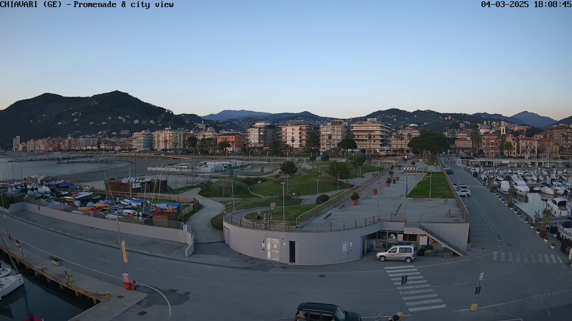 An aerial view of a harbor promenade and city, under a clear twilight sky, shows a paved area with parked cars, a marina filled with boats, and a grassy park area leading to buildings on a hillside.
