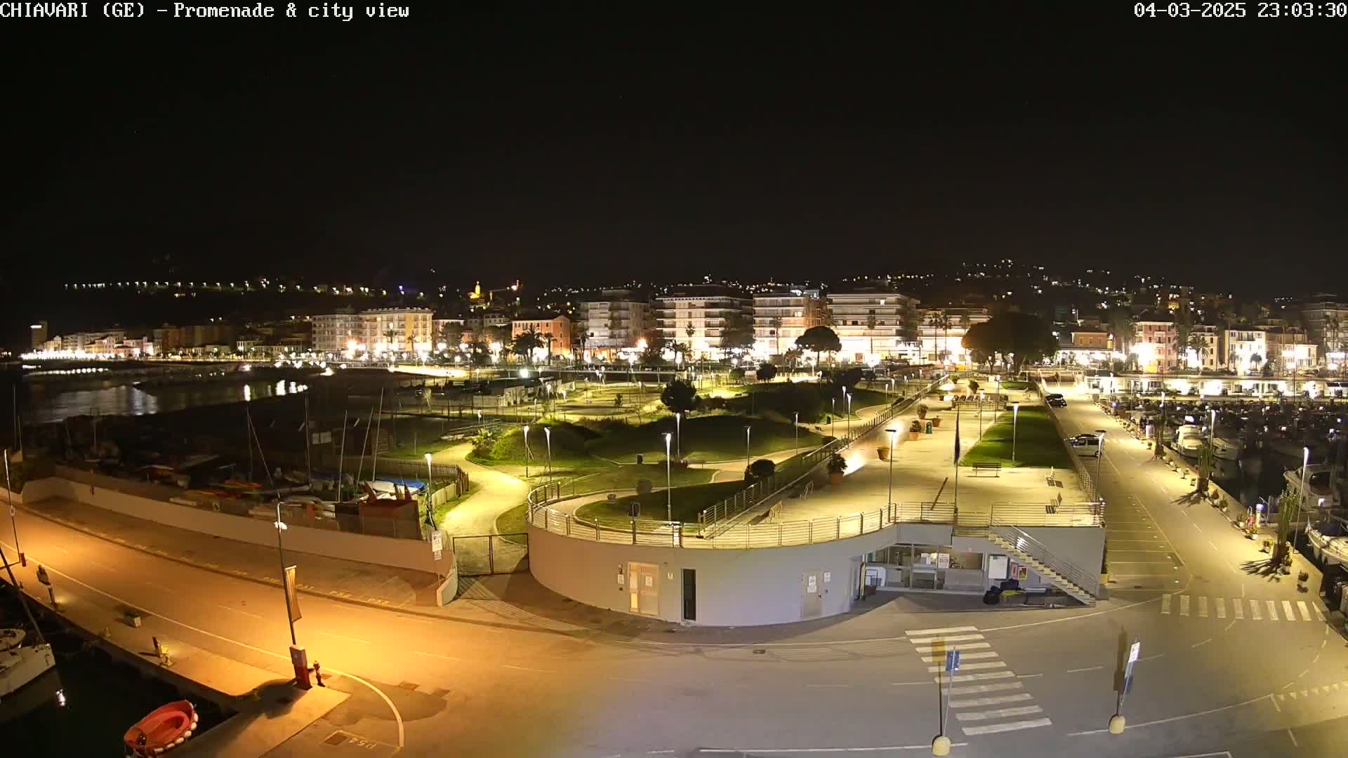 A nighttime, illuminated view of a harbor promenade and city, with boats docked and a park-like area separating the waterfront from buildings.