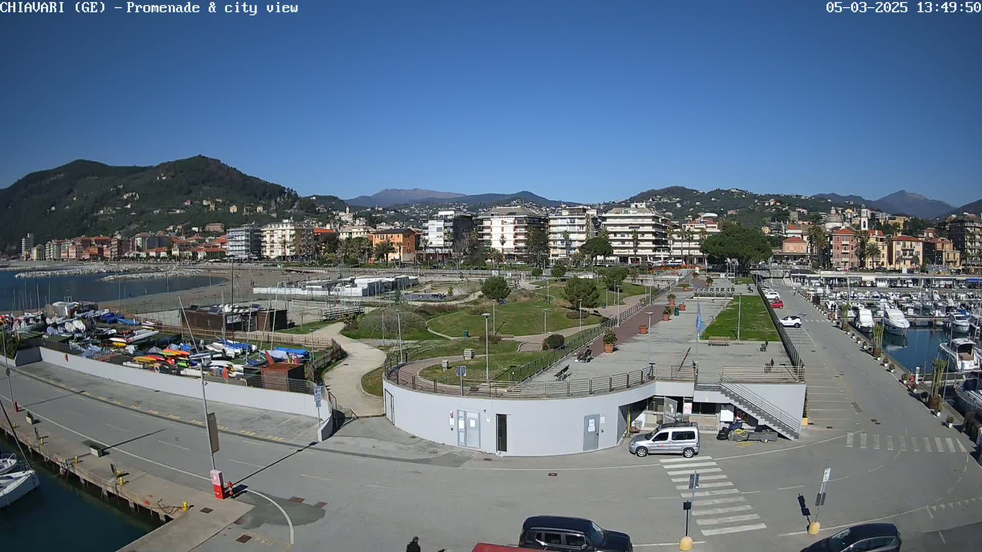 A coastal city scene on a sunny day shows a harbor with boats, a promenade with green spaces, and buildings nestled against a backdrop of mountains.