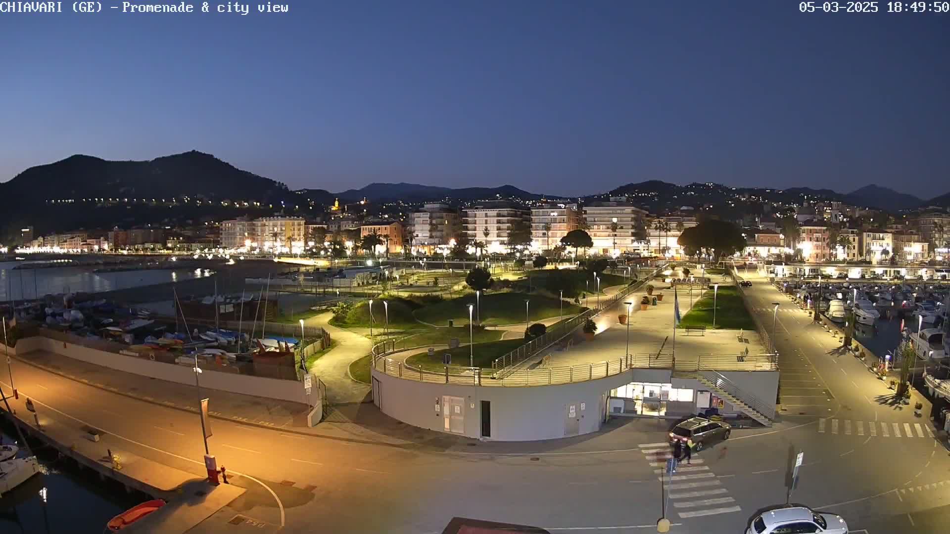 A nighttime view of a coastal town's illuminated promenade, marina, and buildings, under a twilight sky.