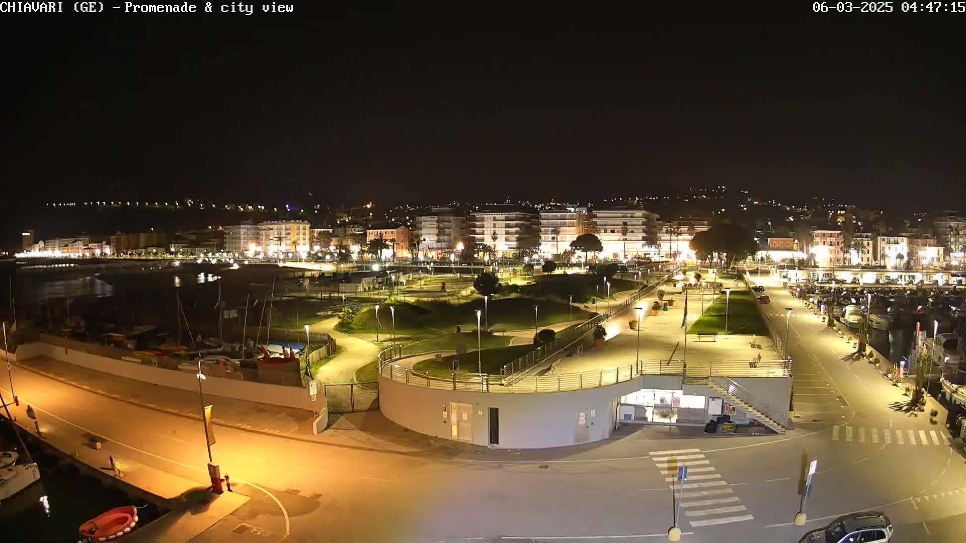 A nighttime view of a brightly lit waterfront promenade and city, with a marina and a park visible.