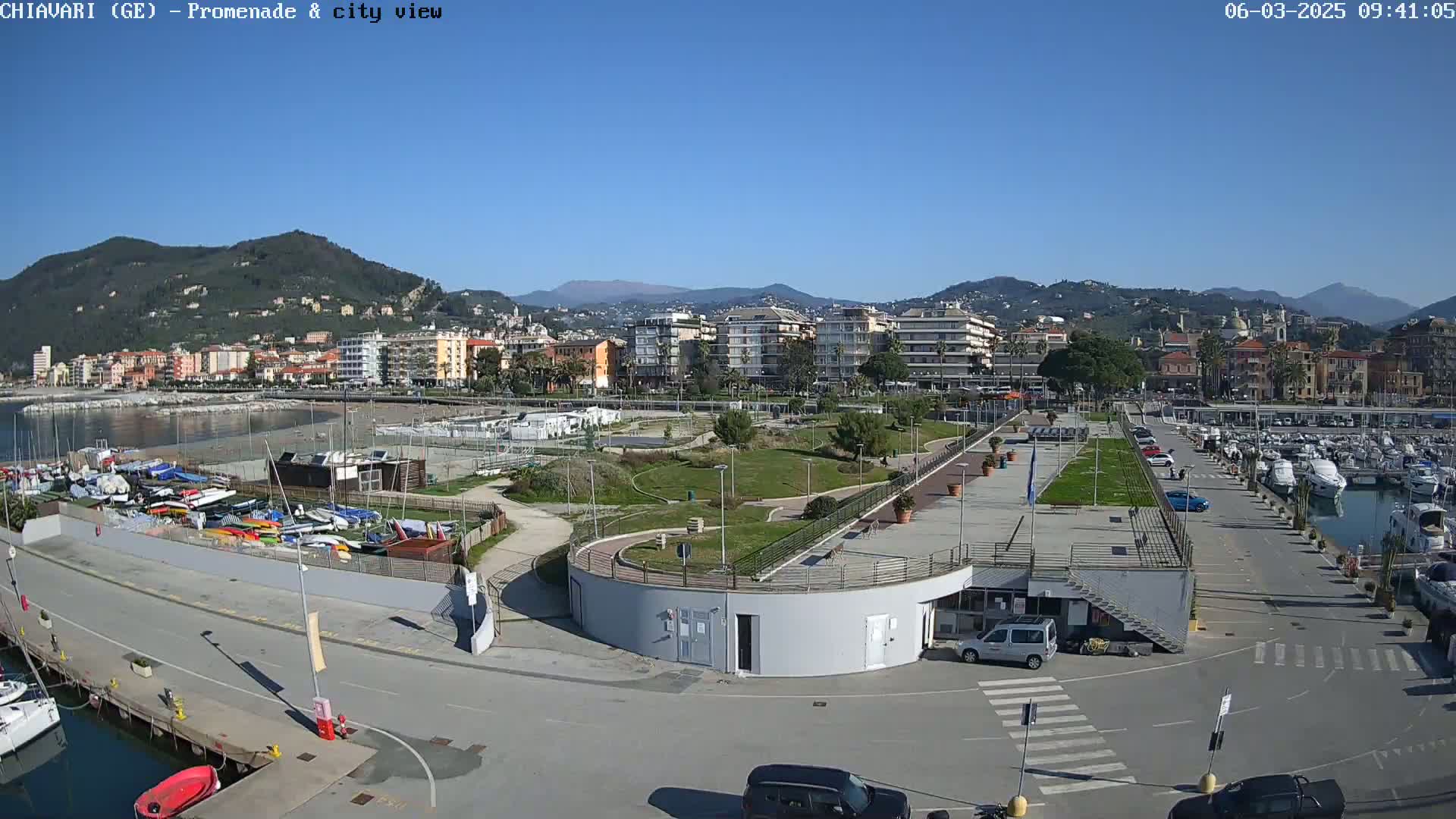 A sunny day overlooking a harbor filled with boats, a paved promenade, a grassy park, and a coastal town nestled against a mountain range.
