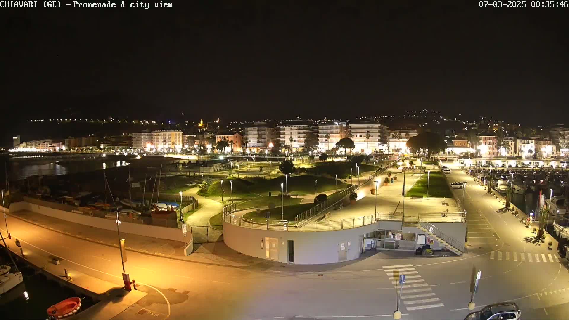 A nighttime view of a brightly lit waterfront promenade and city, with boats moored in the harbor.