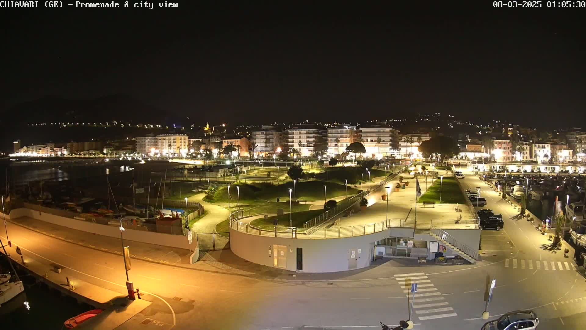 A nighttime view of a city's waterfront promenade and harbor, showing illuminated buildings, landscaped areas, parked cars, and boats in the marina.