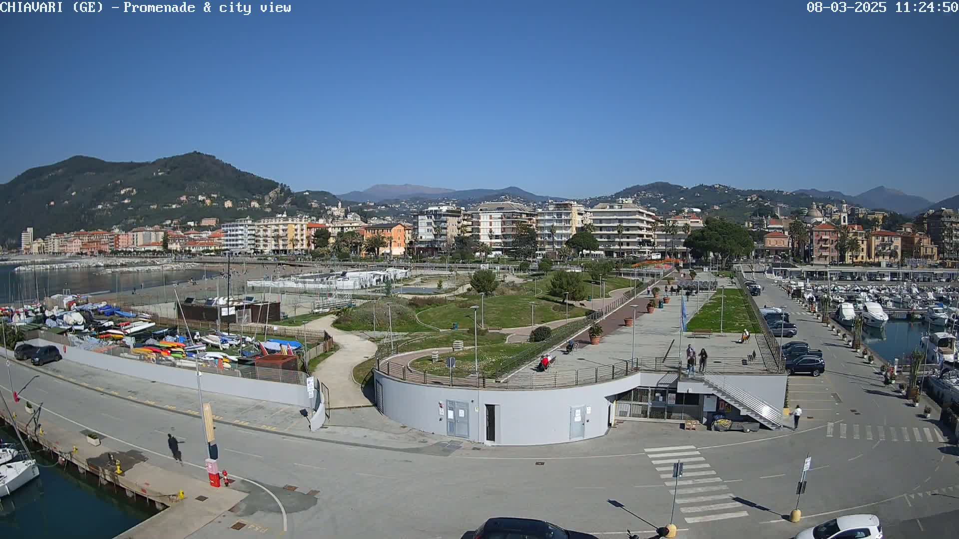 A sunny day reveals a coastal town's promenade, marina, and park, with mountains in the background.