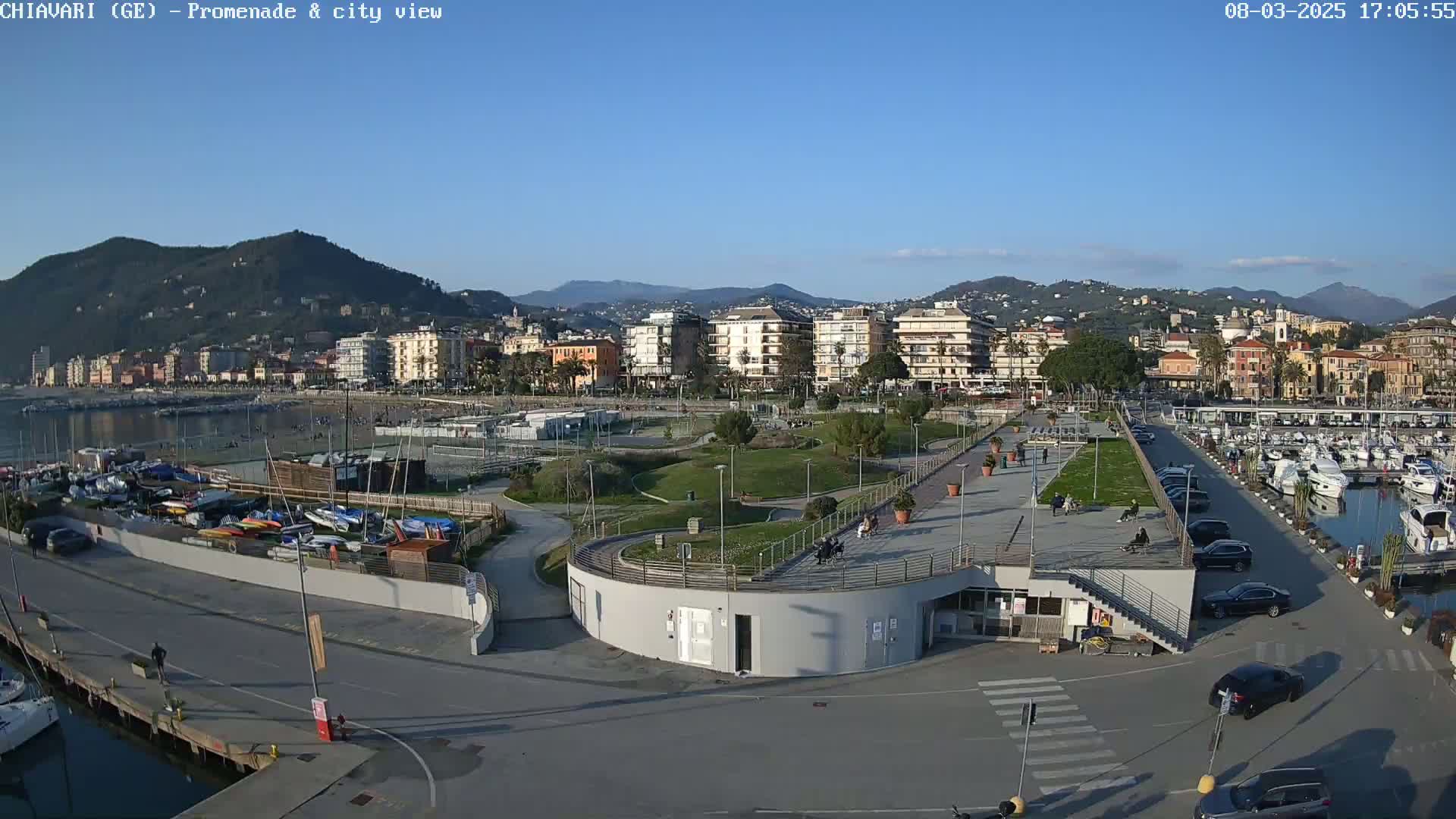 A waterfront promenade with a marina, a park, and a town nestled against a mountain range under a clear blue sky.