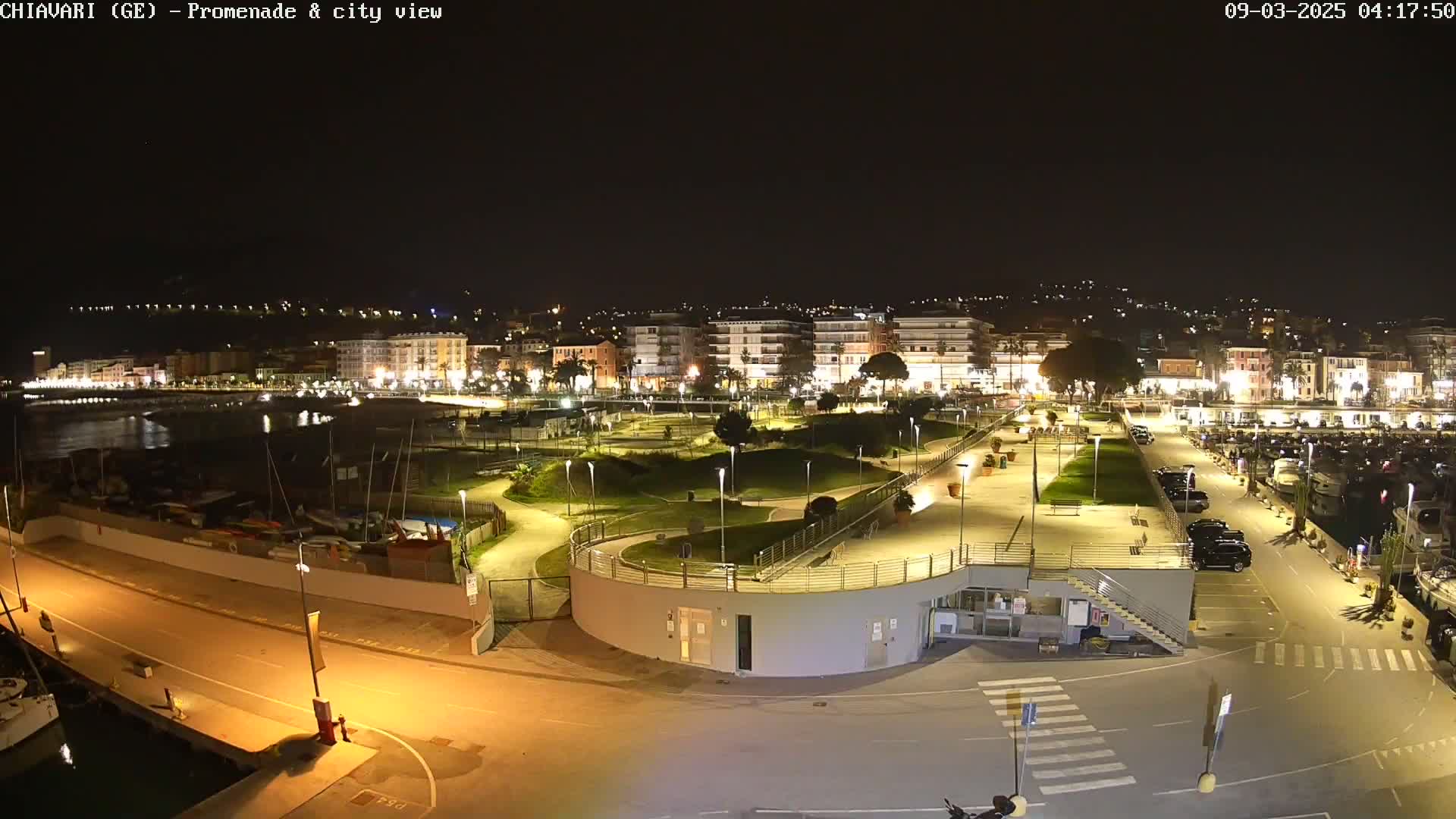 A nighttime aerial view of a city's waterfront promenade and harbor, illuminated by artificial lights, showing a calm body of water and parked cars.