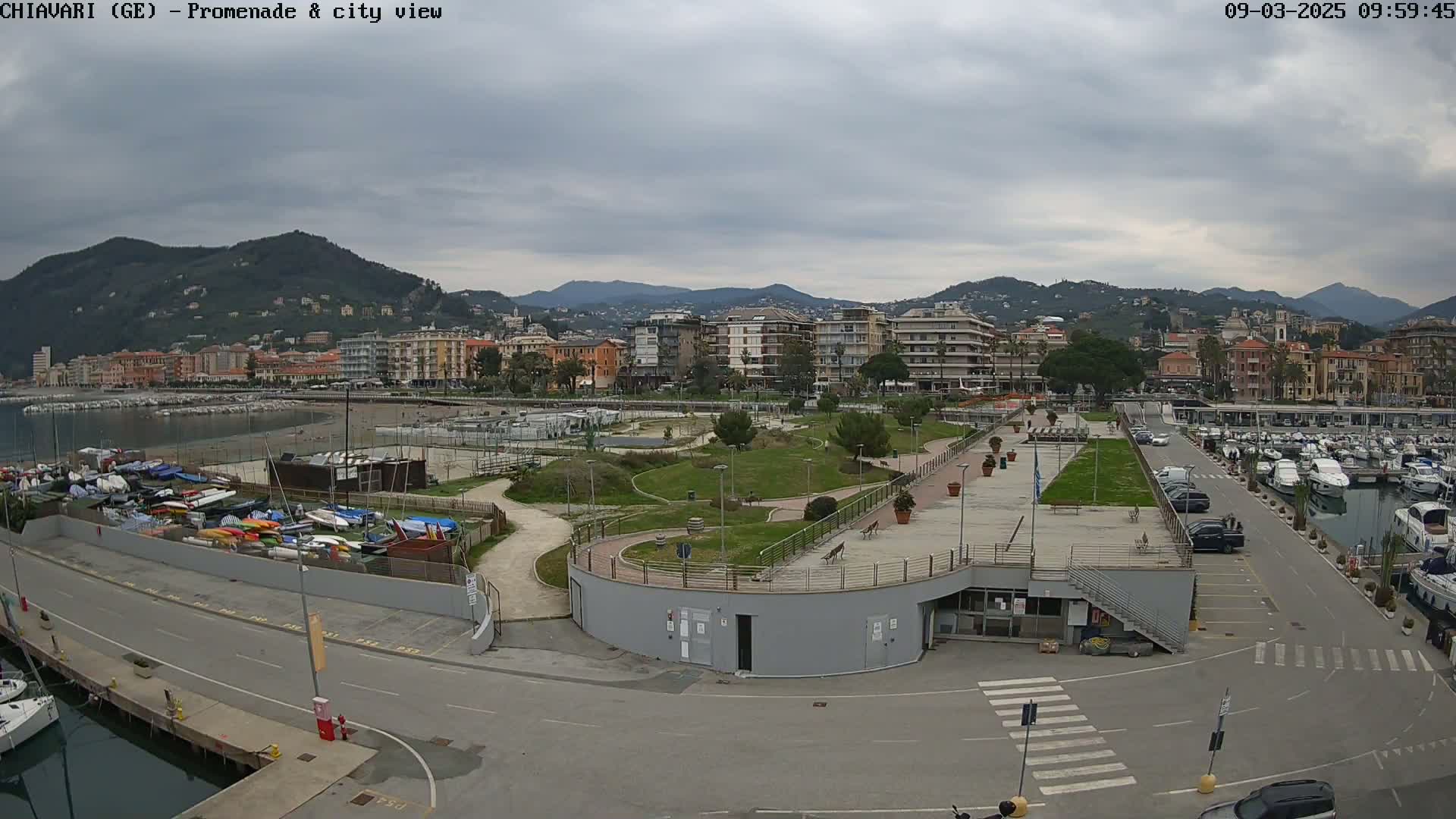 An overcast day reveals a coastal city scene with a promenade, a marina filled with boats, and a park-like area separating the waterfront from buildings nestled against a mountain range.