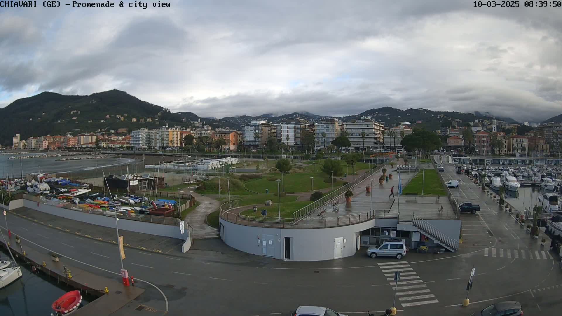 A coastal city scene on an overcast day shows a harbor filled with boats, a promenade, green spaces, and buildings nestled against a hillside.