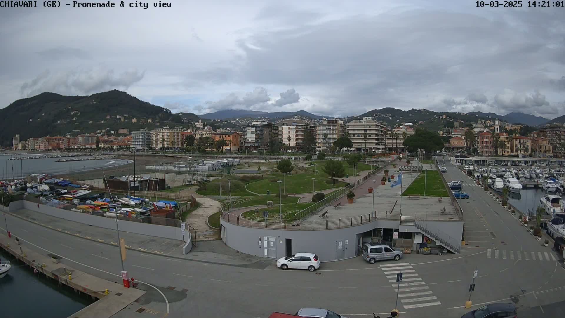 A coastal city with a promenade, marina, and park is visible under a cloudy sky.