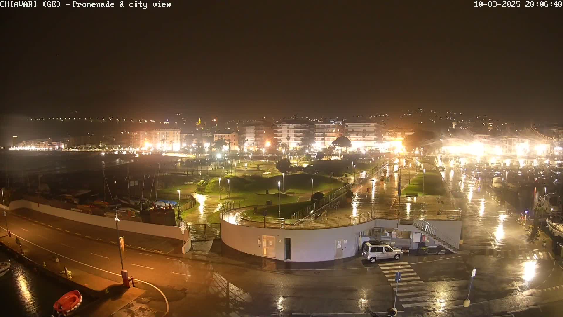 A rain-slicked waterfront promenade and city at night, illuminated by streetlights, with a small vehicle parked near a building.