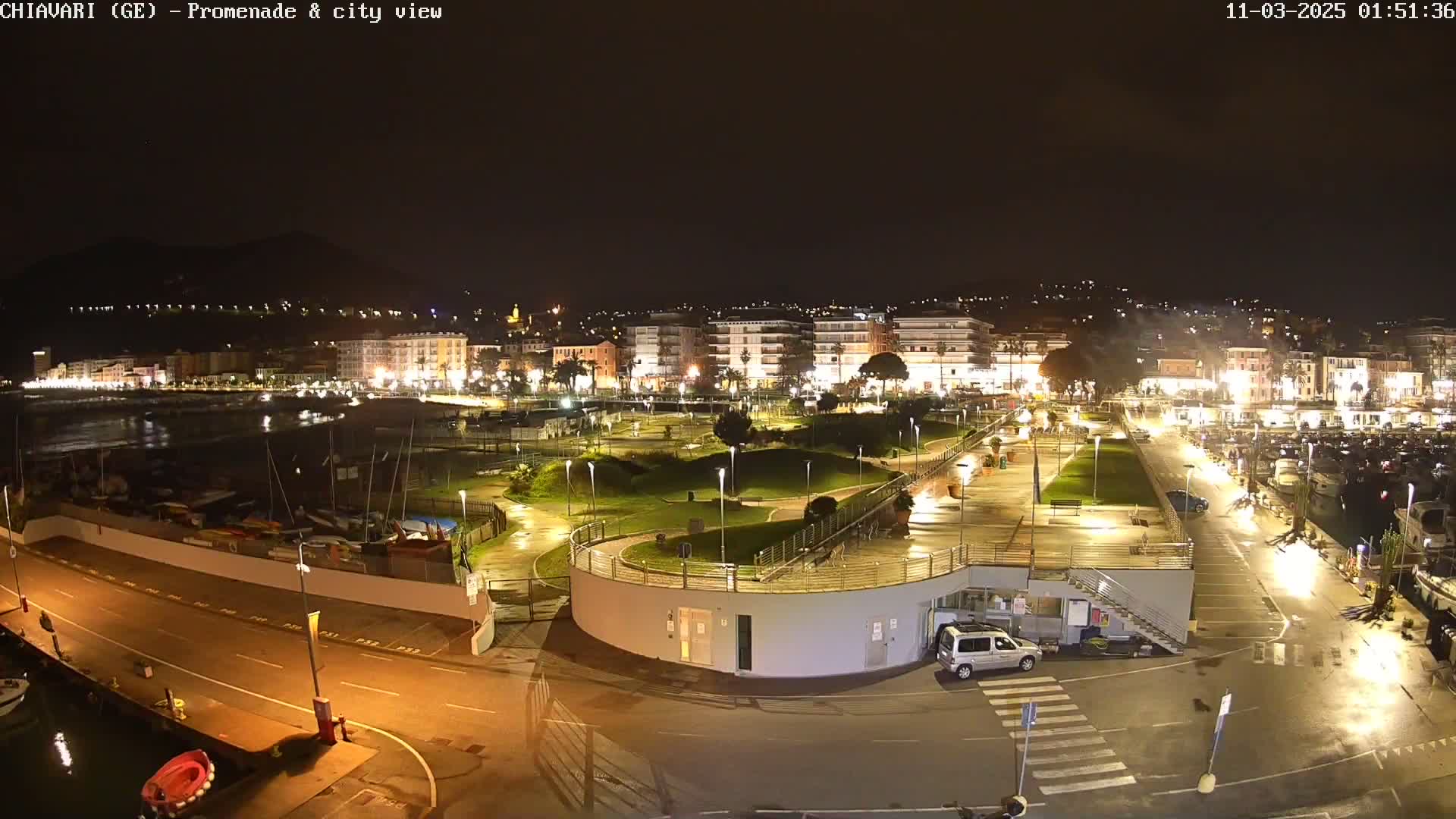 A nighttime view of a city's waterfront promenade and harbor, illuminated by streetlights, showing a calm body of water and numerous boats docked.