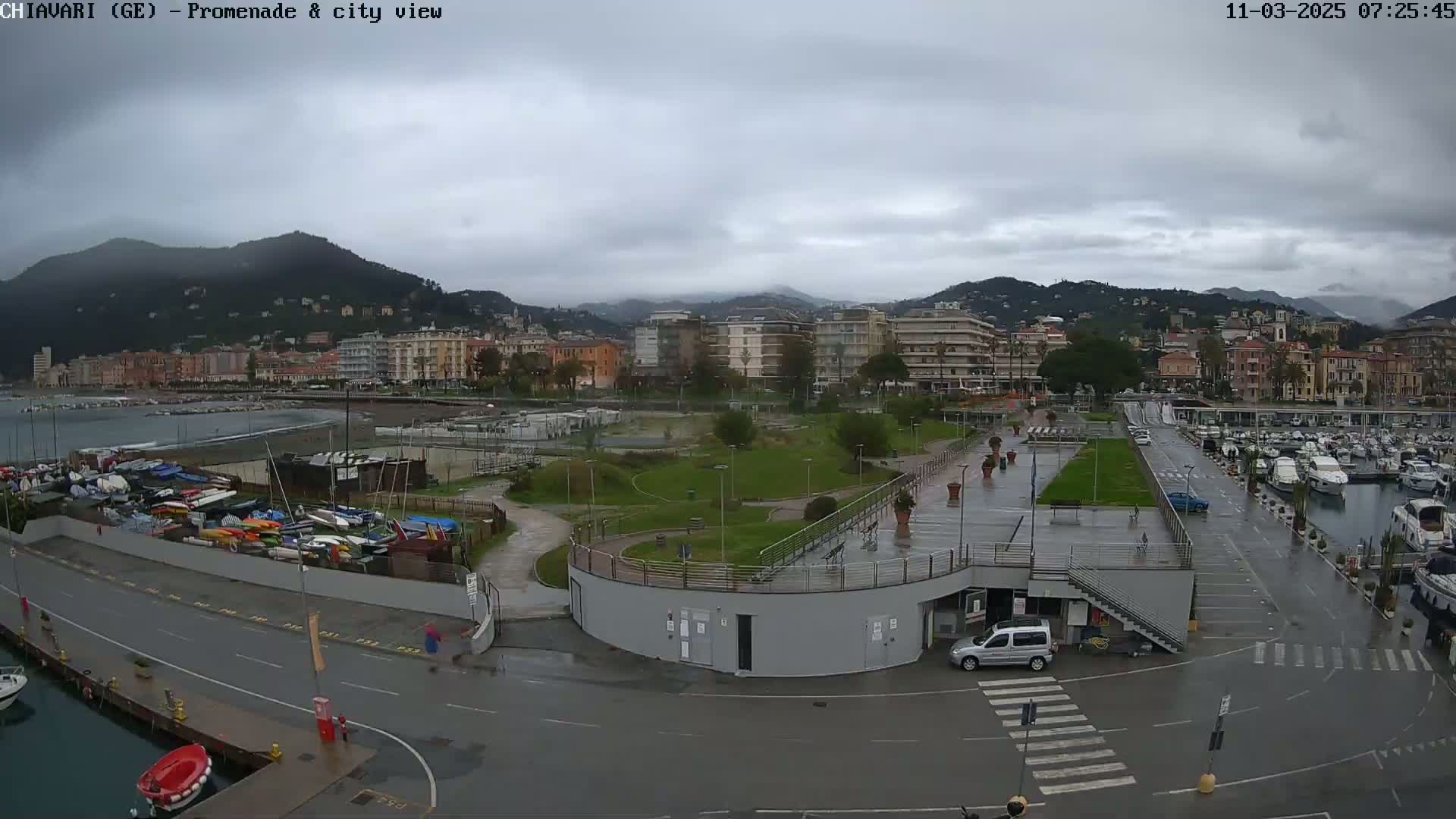 A waterfront city scene under a cloudy sky shows a marina filled with boats, a promenade with a park, and buildings on a hillside in the background.